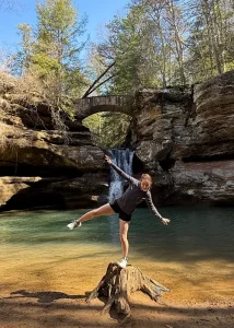 Rachel hiking and standing on a stump in front a waterfall