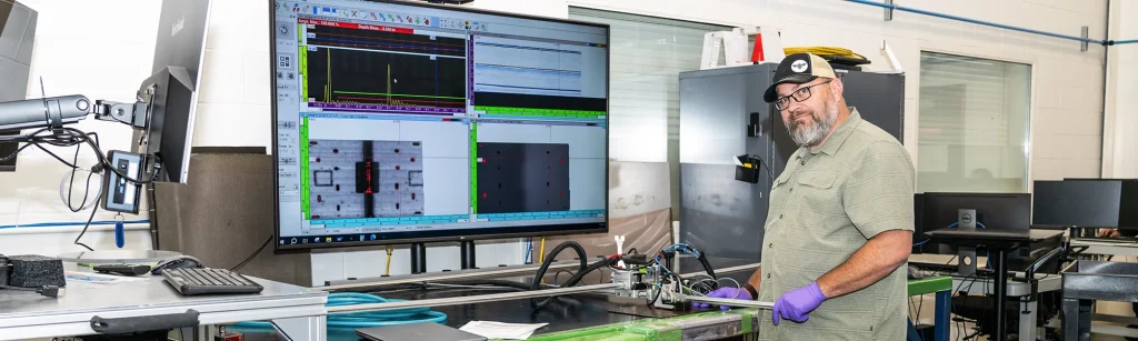 Thomas, NDI Technician, runs phased array NDI equipment. There are waves on the TV screen behind him.